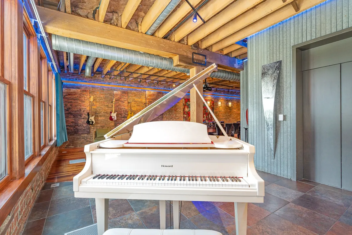 A vibrant living space featuring a grand piano, exposed brick walls, guitars on display, and an elevated ceiling with beams. The arrangement invites musical performances and group gatherings, with an artistic ambiance throughout.