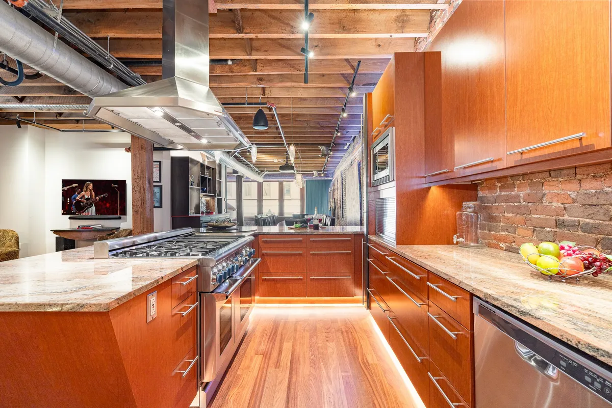 This image features a contemporary kitchen space with a central island, sleek stainless steel appliances, granite countertops, and an exposed brick wall. The area is set up for both cooking and entertaining, with bar seating at the island and warm ambient lighting, capturing the energy of shared meals and gatherings.