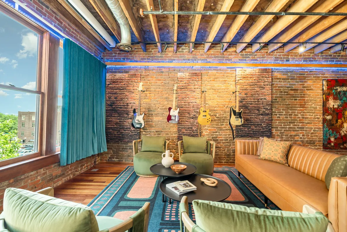 A cozy, creative living area with an exposed brick wall, hanging guitars, a colorful area rug, and stylish seating. The space radiates a relaxed, artistic vibe, perfect for casual conversations or impromptu music sessions.