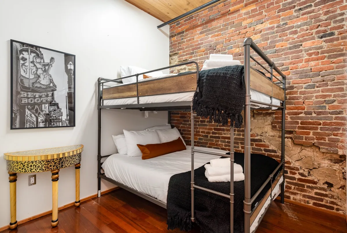 A comfortable bedroom with a queen-over-queen bunk bed, exposed brick wall, decorative table, and framed artwork, creating a space ideal for children or friends. The room feels casual, cozy, and perfect for group stays.