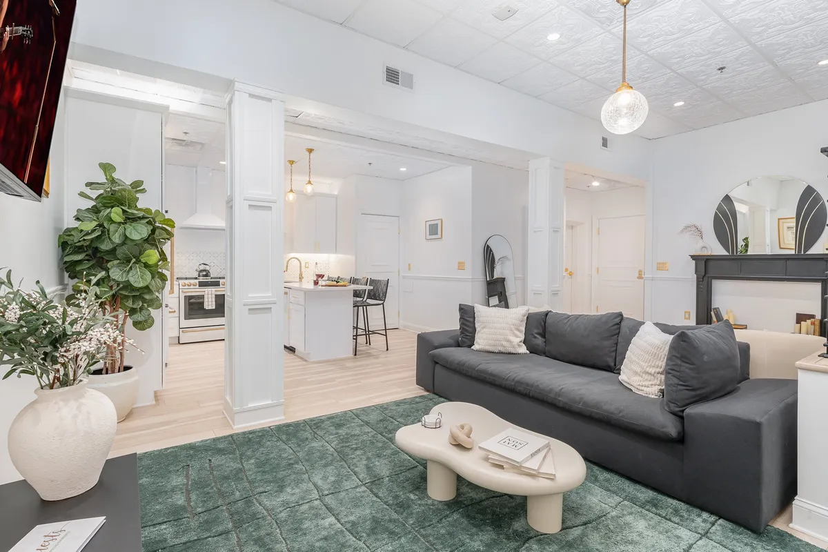A contemporary and cozy living space featuring a sofa, coffee table, decorative plants, kitchen area, and a striking mirror. The open layout hints at the flexibility of the dual-unit configuration, inviting relaxation and entertaining. The kitchen is visible, emphasizing the convenience of having separate social and cooking spaces within each unit.